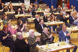 Mehrere Personen sitzen an Tischen in einem Saal und klatschen