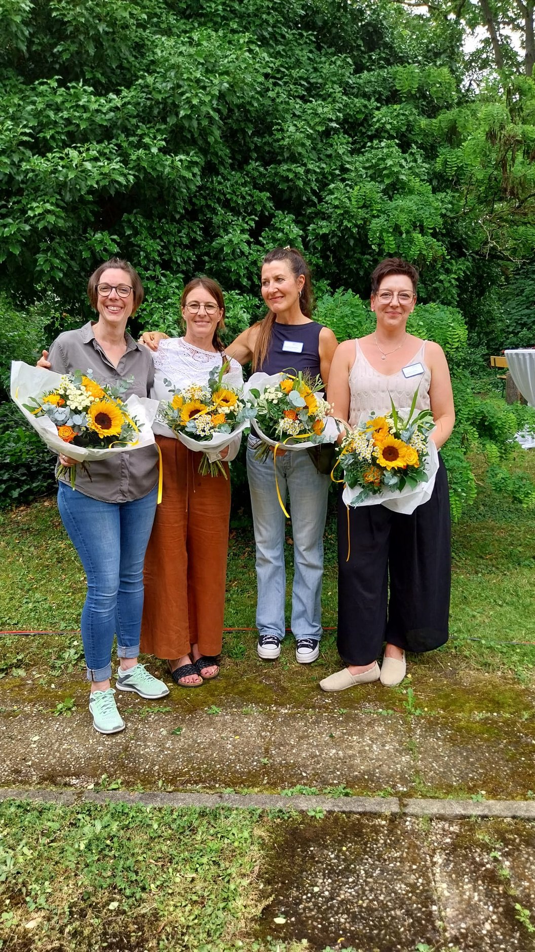 Vier Frauen stehen nebeneinander im Freien, jede hält einen Blumenstrauß mit Sonnenblumen, im Hintergrund dichter grüner Bewuchs