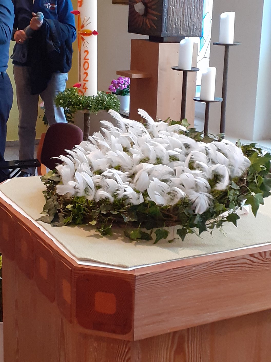 Kranz mit weißen Federn und grünen Blättern auf einem hölzernen Altar in einem Kirchenraum.