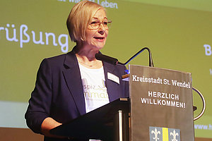 Frau spricht am Rednerpult bei einer Veranstaltung in der Kreisstadt St. Wendel, im Hintergrund Banner.