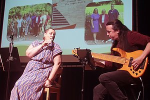 Zwei Personen auf einer Bühne, eine sitzt auf einem Stuhl und hält ein Mikrofon, die andere spielt E-Bass, im Hintergrund eine Präsentation mit dem Titel 'Generationenwechsel 2024' und zwei Fotos von Gruppen.