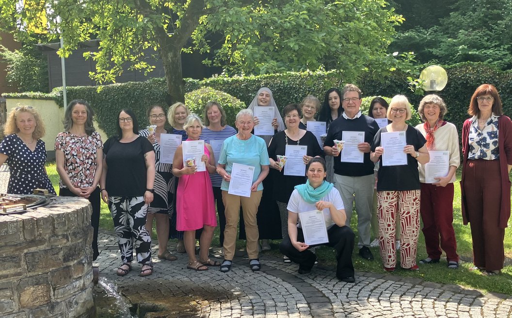 Gruppe von Erwachsenen steht im Garten, einige halten Urkunden in der Hand, umgeben von Bäumen und Hecken.