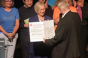 Überreichung eines Schecks über 5000 Euro an eine Frau auf der Bühne, umgeben von applaudierenden Anwesenden.
