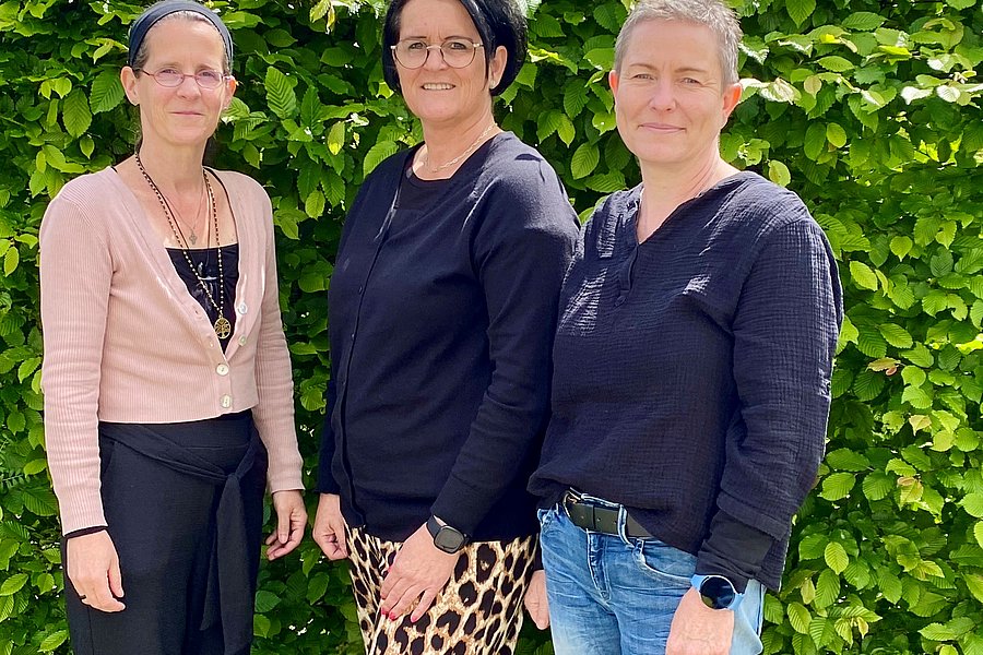 Drei Frauen stehen nebeneinander vor einer grünen Hecke, tragen unterschiedliche Kleidung: rosa Cardigan mit schwarzem Rock, schwarzes Oberteil mit Leopardenmuster-Rock, schwarzes Oberteil mit blauer Jeans.