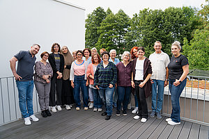 Gruppe von Menschen unterschiedlichen Alters steht auf einer Dachterrasse vor Bäumen und einer weißen Wand