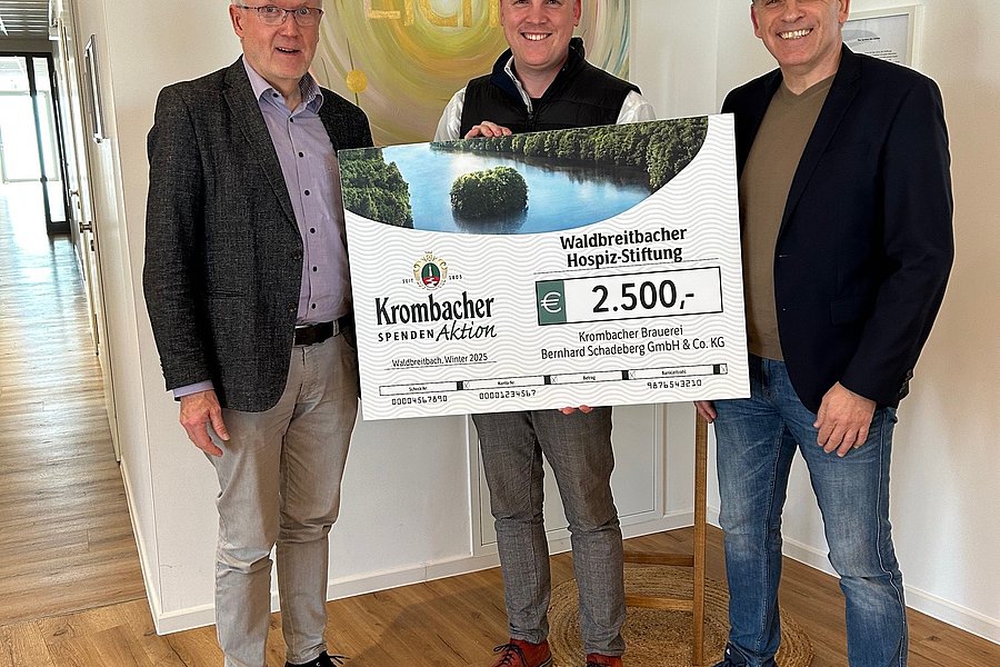 Drei Männer stehen in einem Raum und halten gemeinsam einen großen symbolischen Scheck über 2.500 Euro von Krombacher für den Naturschutzbund Region Südwestfalen