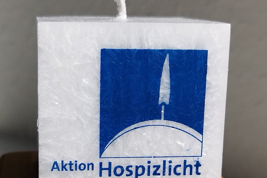 Weiße quadratische Kerze mit blauem Logo und Text 'Aktion Hospizlicht In Würde leben – bis zuletzt' auf Holzuntergrund