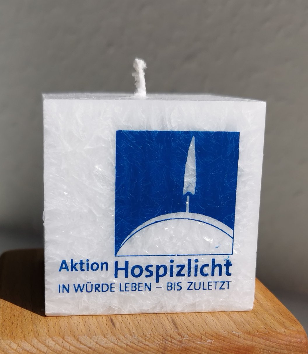 Weiße quadratische Kerze mit blauem Logo und Text 'Aktion Hospizlicht In Würde leben – bis zuletzt' auf Holzuntergrund