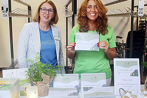 Zwei Frauen an einem Stand mit Kalendaren und Pflanzen, lächeln in die Kamera.