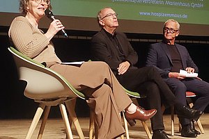 Drei sitzende Personen auf einer Bühne, eine spricht in ein Mikrofon, im Hintergrund eine grüne Projektionsfläche mit Text