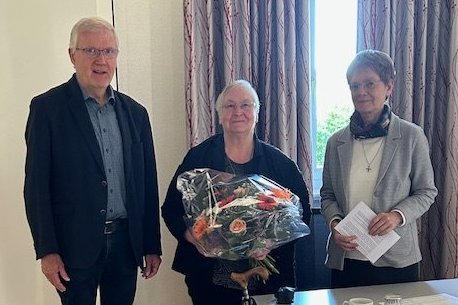 Drei Personen stehen in einem Raum, eine Person hält einen Blumenstrauß, auf dem Tisch liegen Tassen und Papiere