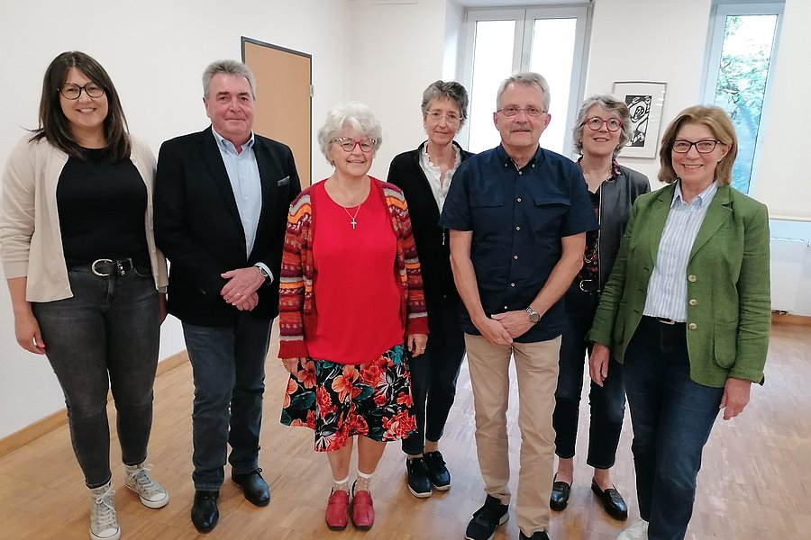 Gruppe von sieben Personen in einem hellen Raum, unterschiedliche Kleidung, stehend, einige tragen Jacken oder Blazer.