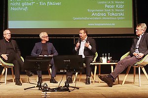 Podiumsdiskussion mit vier Männern auf Stühlen vor einer grünen Präsentationsfolie mit Text zu Hospiz Emmaus und Namen von Referenten