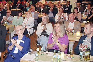 Menschen applaudieren bei einer Veranstaltung, einige sitzen an Tischen mit Getränke.