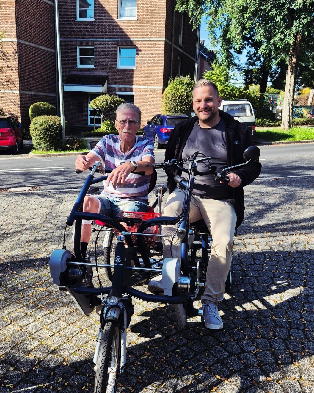 Zwei Personen sitzen auf einem dreirädrigen Tandemfahrrad auf einer gepflasterten Straße vor einem Backsteingebäude.