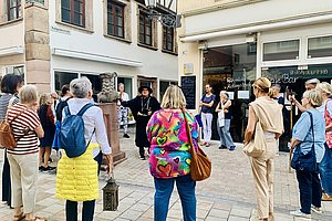 Gruppe von Menschen steht in einem Innenhof und hört einem Mann in historischer Kleidung zu, der eine Laterne hält und spricht