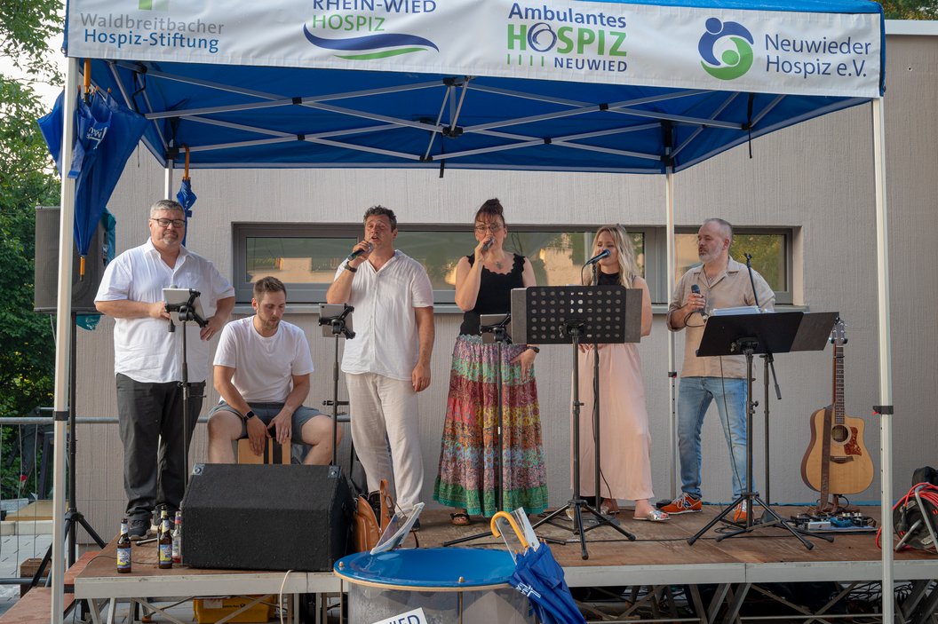 Sechs Personen auf einer Bühne unter einem blauen Zelt mit Bannern von Hospiz-Stiftungen, sie singen und spielen Musik, eine Gitarre steht rechts
