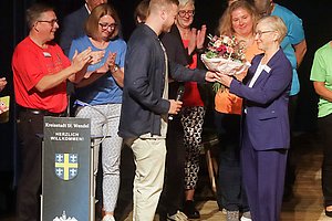 Person überreicht einer anderen Person auf einer Bühne einen Blumenstrauß, im Hintergrund applaudieren mehrere Personen.