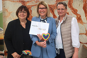 Drei Personen stehen vor einer Wand mit rotem Muster, die mittlere Person hält ein Schild mit der Aufschrift 'Hospiz für ... VIELFALT' und ein Herz in Regenbogenfarben, die linke Person hält ein Regenbogen-Symbol.