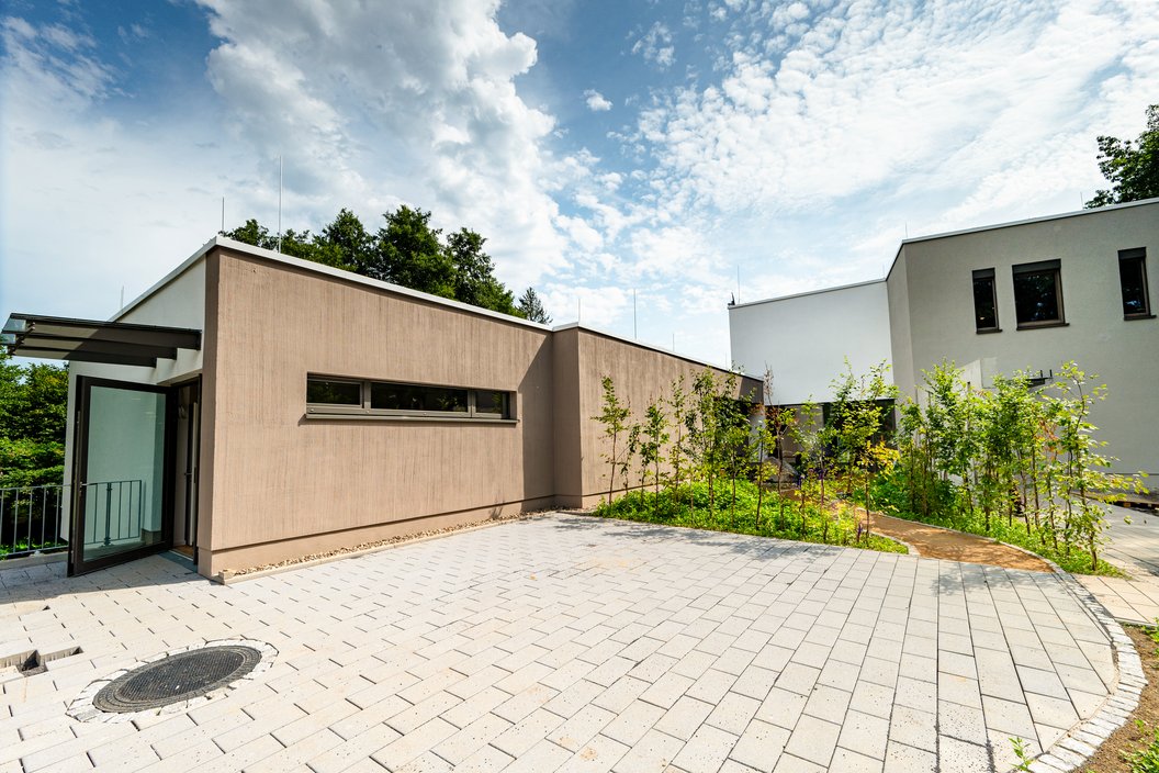 Modernes, eingeschossiges Gebäude mit flachem Dach, hellbrauner Fassade und großen Fenstern, umgeben von gepflastertem Hof und Büschen.
