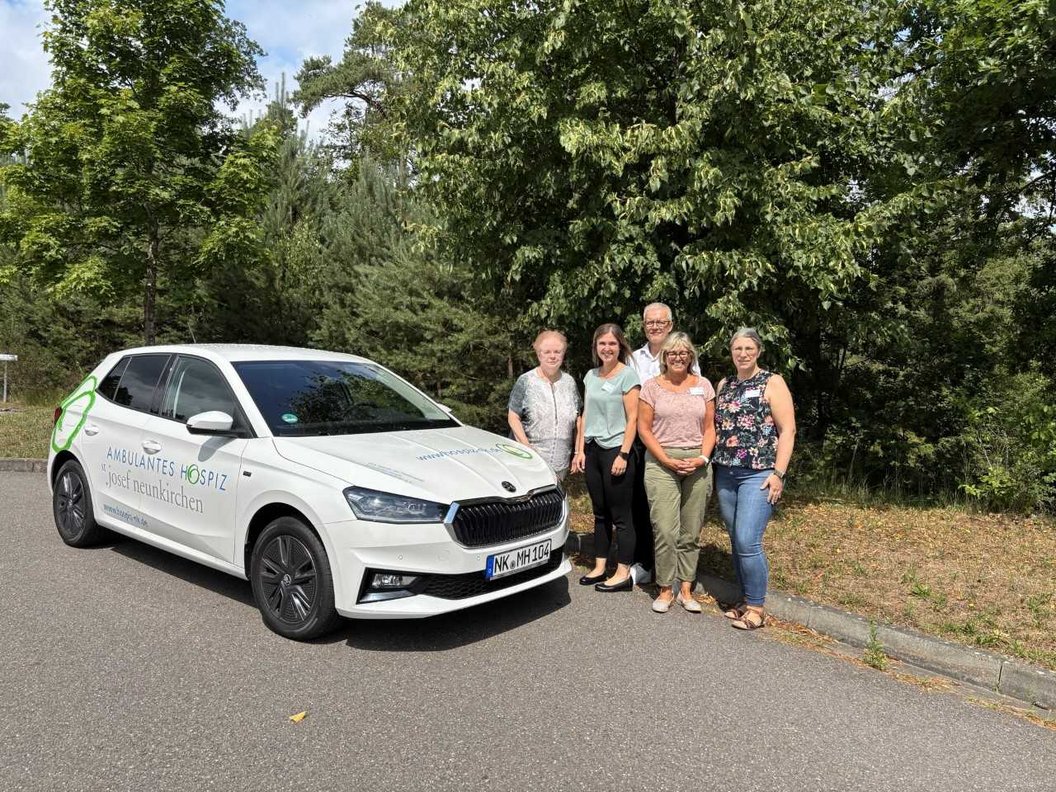 Weißes Auto mit der Aufschrift 'AKB Diakonie Hospiz' steht auf einer Straße neben fünf Personen vor Bäumen.