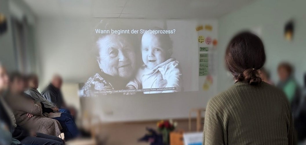 Person betrachtet eine Projektion mit einer alten Frau und einem Baby, darüber der Text 'Wann beginnt der Sterbeprozess?' in einem Raum mit mehreren sitzenden Personen