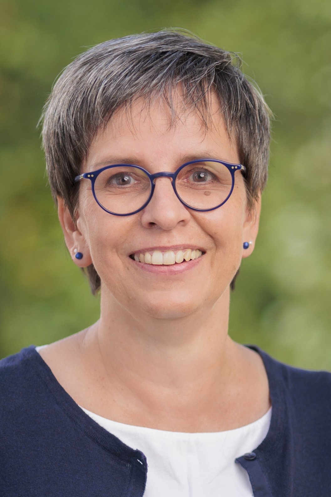 Hiltrud Sachs mit kurzem, grauem Haar trägt eine dunkelblaue Strickjacke über einem weißen Oberteil vor unscharf grünem Hintergrund.
