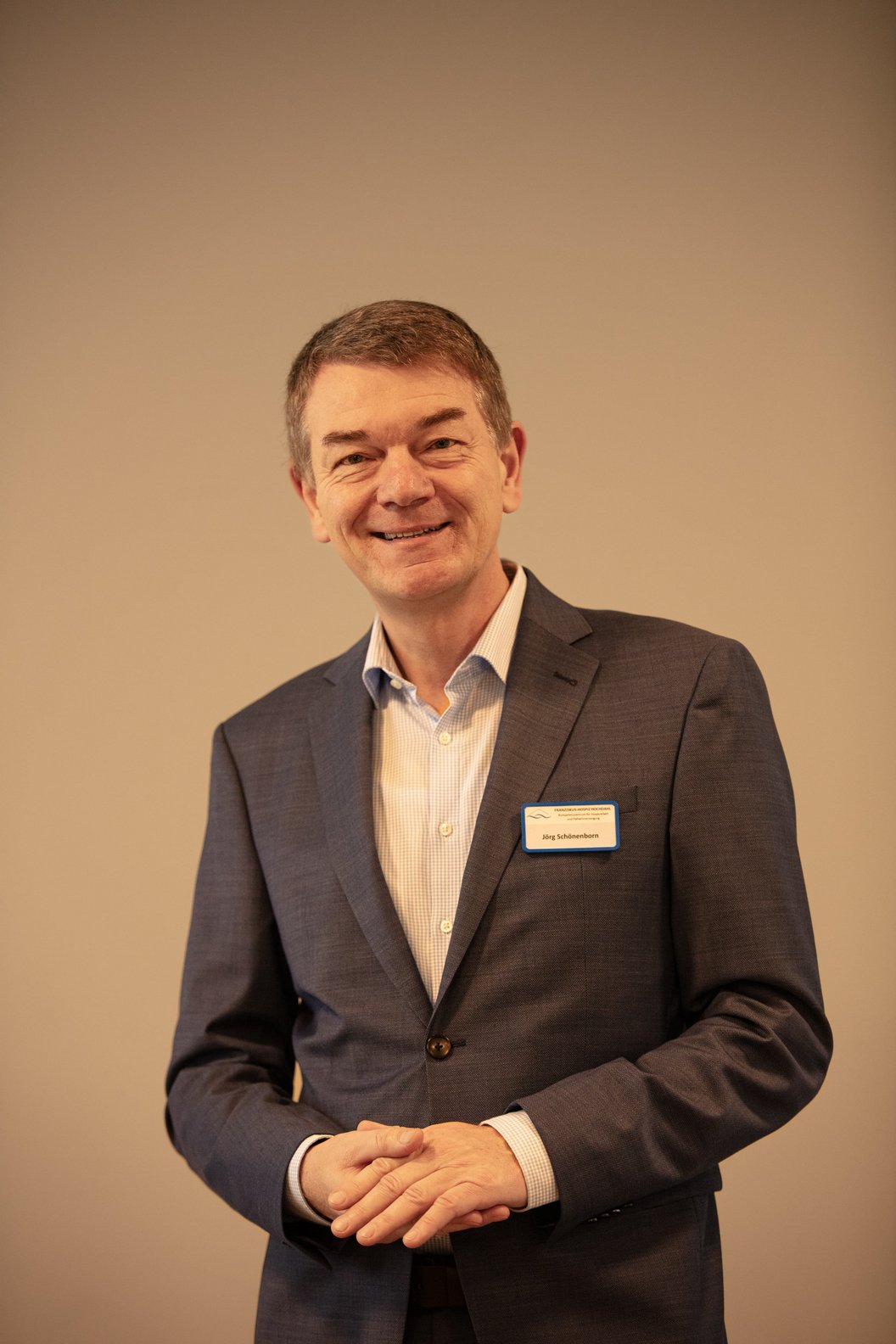 Jörg Schönenborn in dunklem Anzug mit weißem Hemd und Namensschild steht vor beigem Hintergrund und hält die Hände vor dem Bauch.