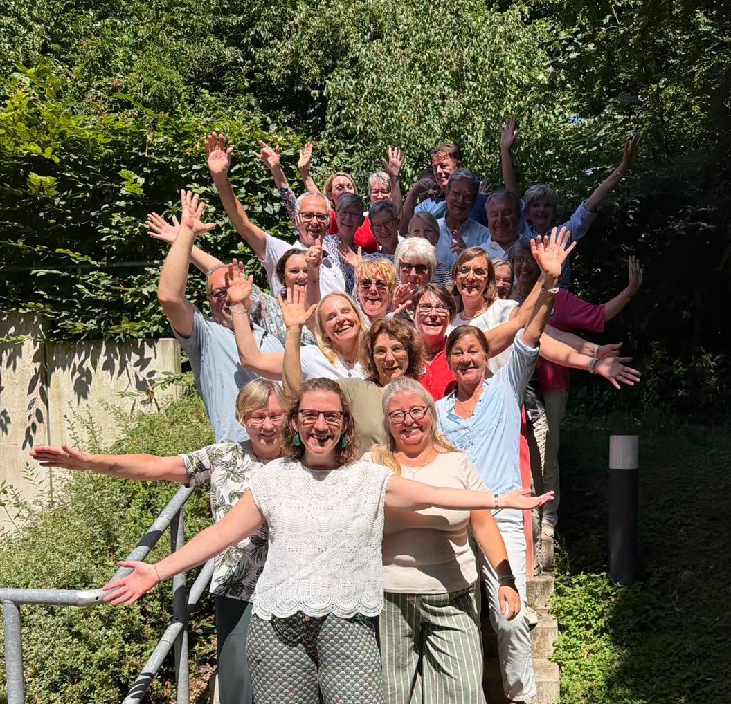 Gruppe von Menschen steht auf einer Treppe, die Arme weit geöffnet, im Freien vor grünem Gebüsch