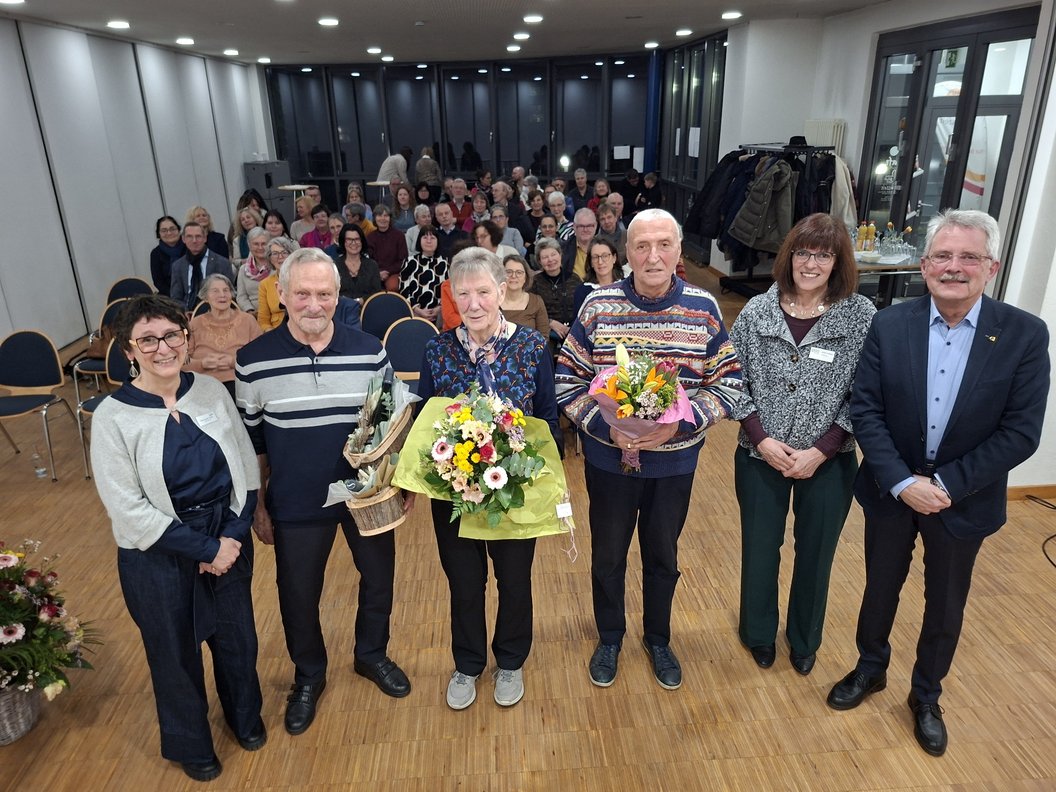 Feierliche Veranstaltung mit mehreren Personen, Blumensträußen und einer großen Gruppe im Hintergrund.