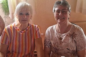 Zwei ältere Frauen sitzen nebeneinander vor einem hellen Vorhang, die linke trägt ein gestreiftes Shirt, die rechte eine Brille und ein gemustertes Shirt