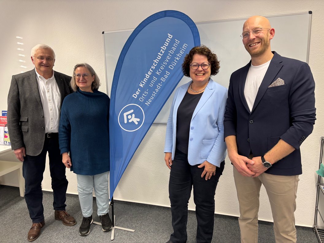 Vier Personen stehen vor einem blauen Werbefahnen mit der Aufschrift 'Der Kinderschutzbund Orts- und Kreisverband Neuss und Bad Dürkheim' in einem Raum mit weißer Wand und Möbeln