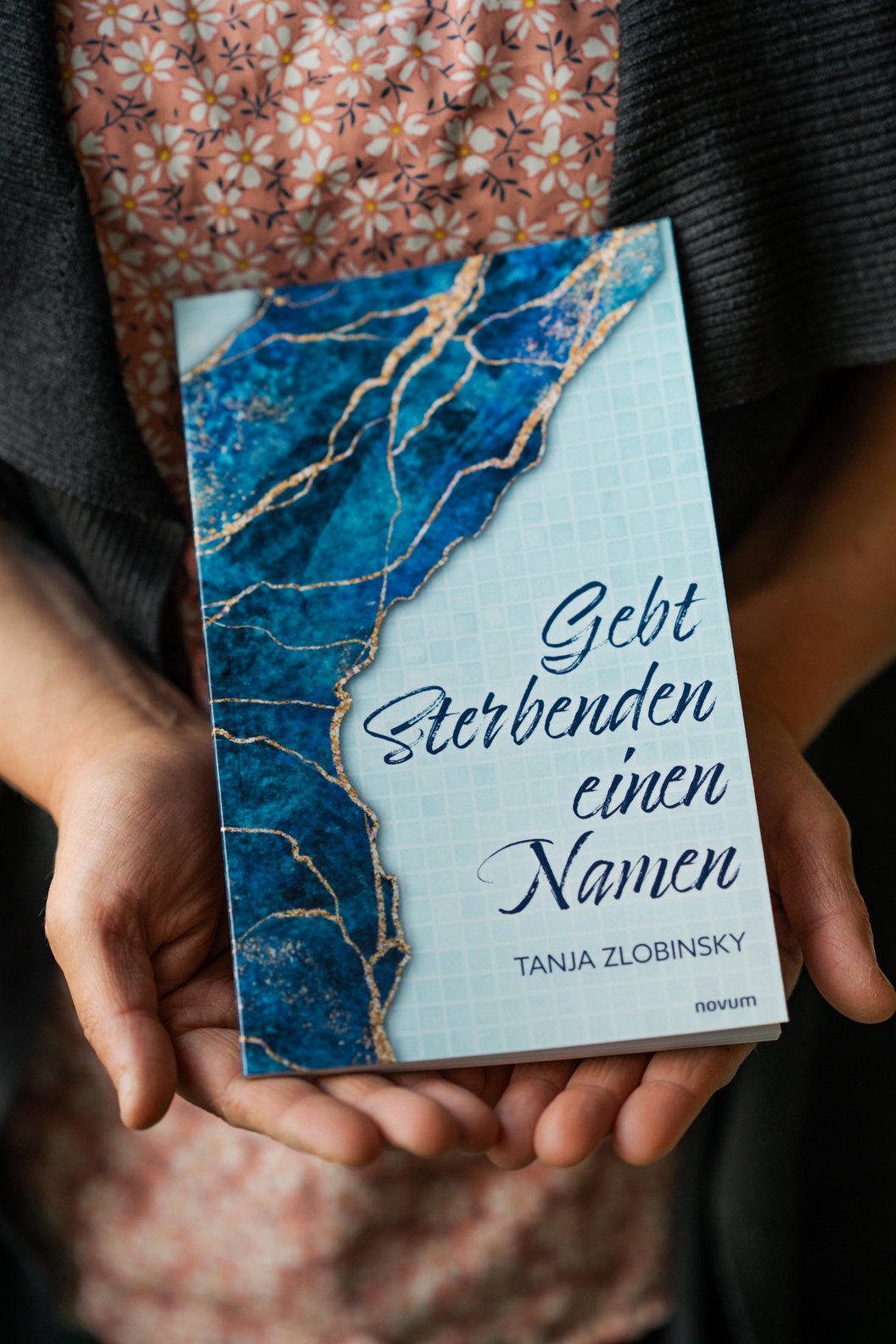 Hände halten ein Buch mit blau-weißem Marmormuster und dem Titel 'Gebt Sterbenden einen Namen' von Tanja Zlobinsky