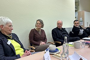 Vier Personen sitzen an einem Tisch mit Tassen, Tellern, einer Wasserflasche und Namenskarten in einem Raum mit hellen Wänden und Schränken.