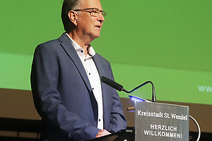 Mann im Anzug steht an einem Rednerpult mit Mikrofon vor grünem Hintergrund