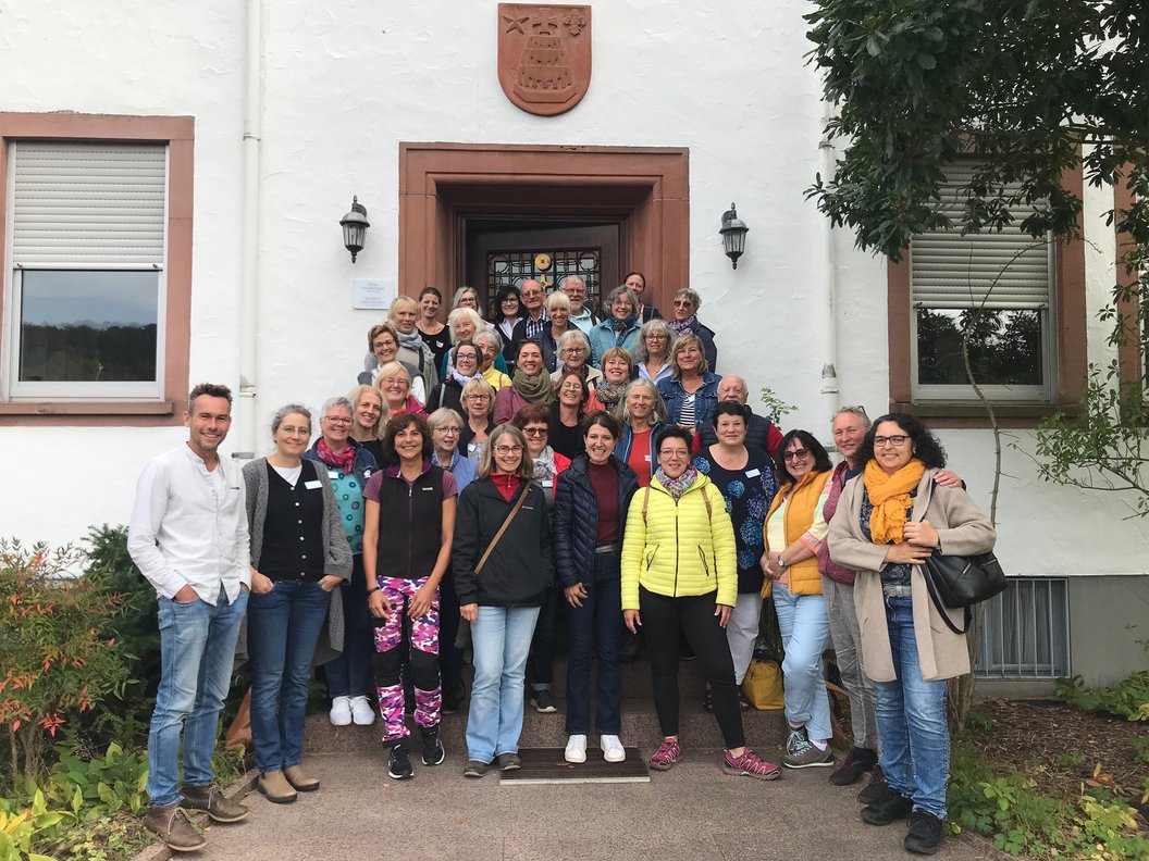 Gruppe von Menschen vor einem Gebäude mit Wappen über dem Eingang, umgeben von Fenstern und Pflanzen.