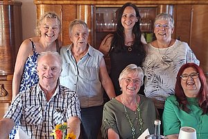 Gruppe von sieben lächelnden Menschen unterschiedlichen Alters in einem Wohnzimmer, drei sitzen an einem Tisch mit Blumen und Getränken, vier stehen dahinter