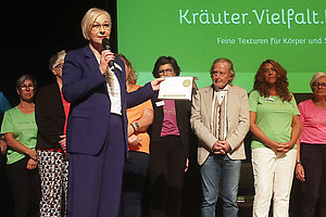 Person in blauem Anzug hält Mikrofon und Urkunde vor einer Gruppe von Menschen auf einer Bühne mit grünem Hintergrund und Text 'Kräuter Vielfalt'