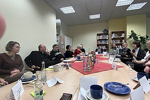 Mehrere Personen sitzen an einem langen Tisch in einem Besprechungsraum, auf dem Tassen, Flaschen und Namenskarten stehen.