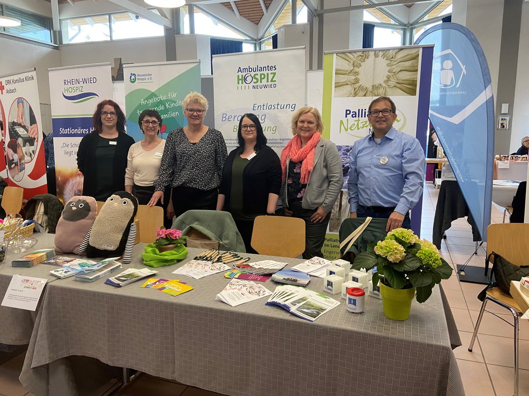 Sechs Personen stehen hinter einem Infostand mit Flyern, Broschüren und Dekoration, im Hintergrund Banner von Hospiz und Palliativnetzwerken