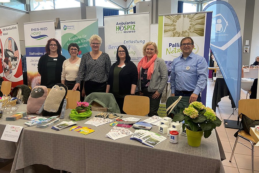 Sechs Personen stehen hinter einem Infostand mit Flyern, Broschüren und Dekoration, im Hintergrund Banner von Hospiz und Palliativnetzwerken
