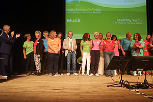 Menschen stehen in einer Reihe auf einer Bühne vor einer grünen Leinwand mit Texten zu Musik und einem Chorprojekt.