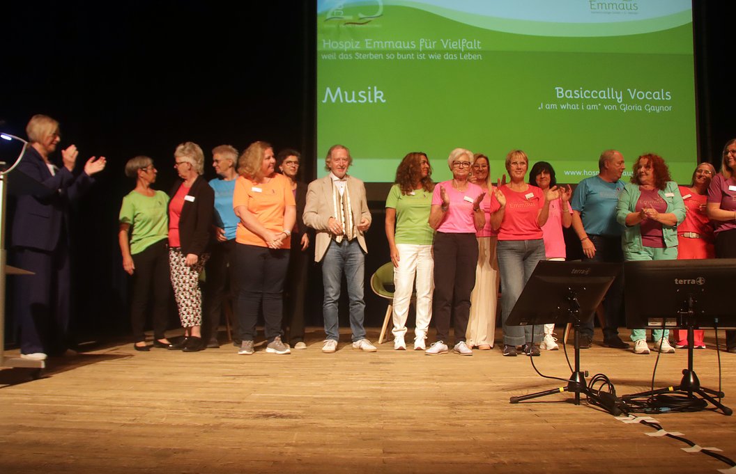 Menschen stehen in einer Reihe auf einer Bühne vor einer grünen Leinwand mit Texten zu Musik und einem Chorprojekt.