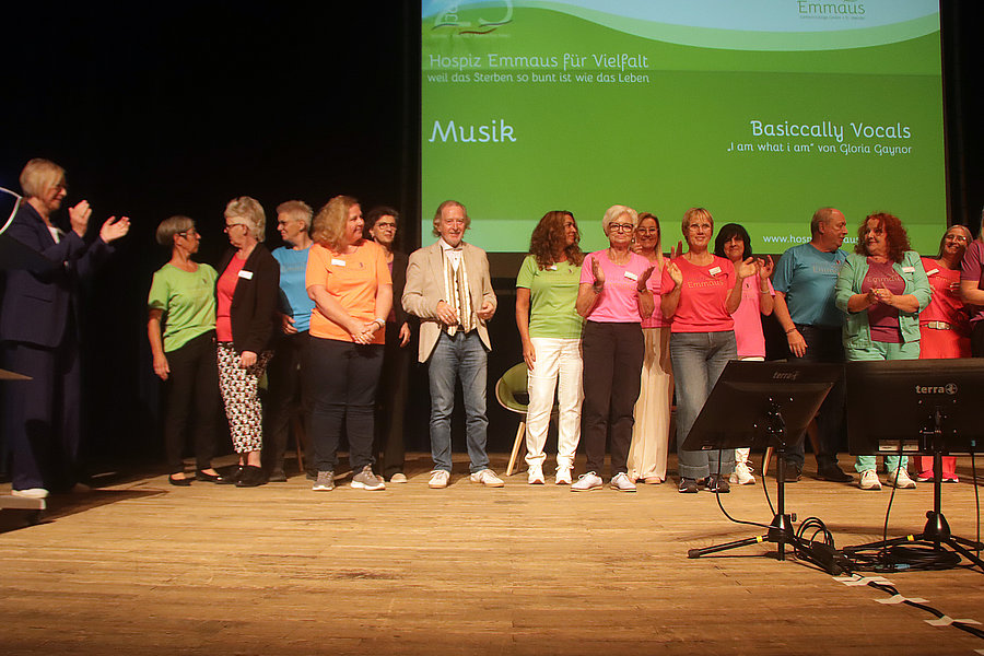 Menschen stehen in einer Reihe auf einer Bühne vor einer grünen Leinwand mit Texten zu Musik und einem Chorprojekt.