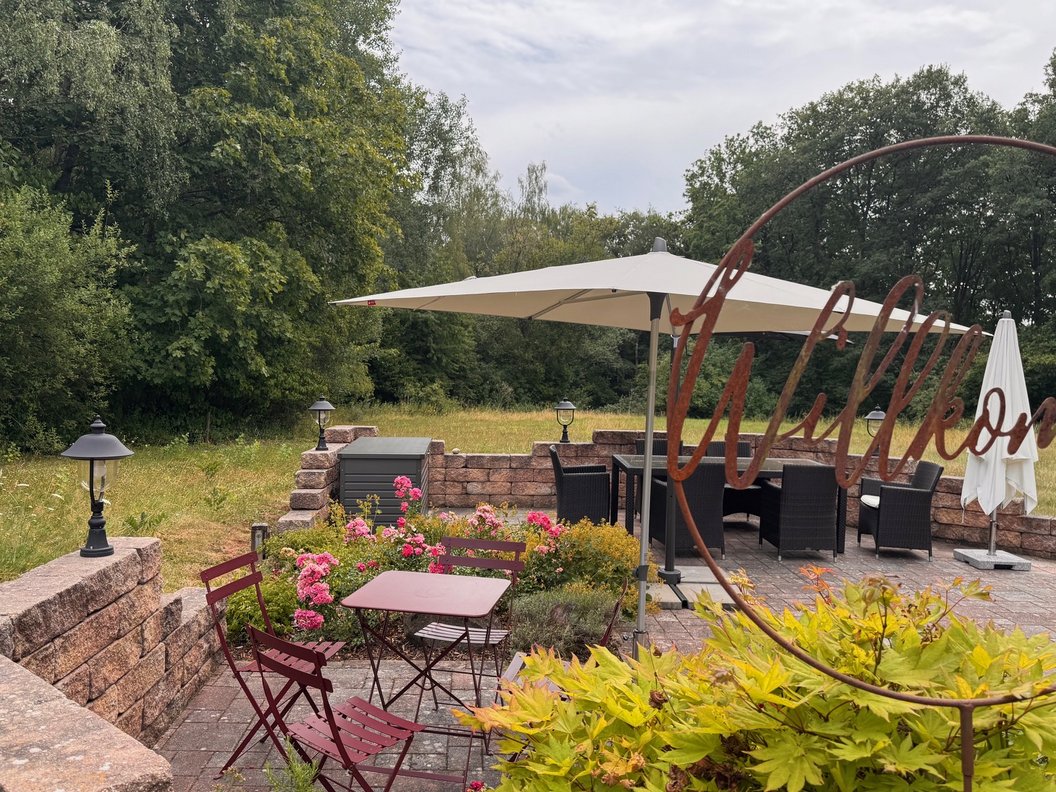 Terrasse mit Tischen, Stühlen, Sonnenschirmen und Pflanzen vor einem Wald