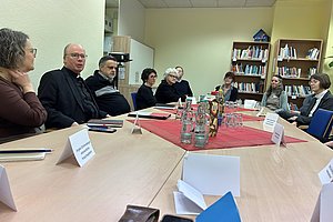 Mehrere Personen sitzen an einem länglichen Tisch mit Namensschildern, Wasserflaschen und Gläsern in einem Raum mit Bücherregal und Schrank.