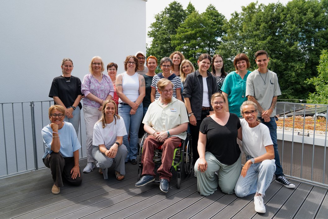 Gruppe von Menschen unterschiedlichen Alters und Geschlechts auf einer Dachterrasse mit Holzplanken, einige sitzen, andere stehen, im Hintergrund Bäume und ein weißes Gebäude