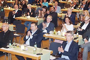 Applaudierende Menschenmenge in einem großen Raum, sitzend an Tischen mit Getränken.