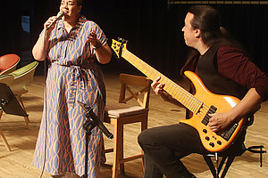 Musikerin singt mit Mikrofon, während ein E-Gitarrist sie im Hintergrund begleitet.