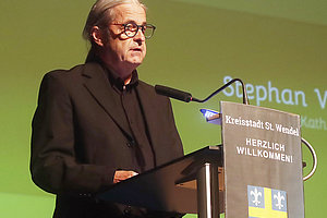 Person mit Brille und dunklem Sakko steht an einem Rednerpult mit dem Schriftzug 'Kreisstadt St. Wendel HERZLICH WILLKOMMEN' vor grünem Hintergrund